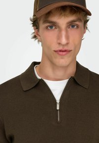 Brązowy dziergany polo na zamek z kołnierzykiem, w połączeniu z białą koszulką pod spodem. Model nosi brązową czapkę z tyłem z siateczki.