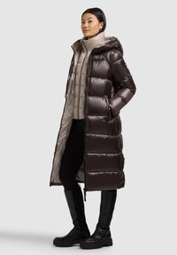 Langer Steppmantel in Dunkelbraun mit glänzendem Finish, mit Kapuze, Reißverschluss und großen gesteppten Partien. Wird über einer hellbeigen Jacke getragen.