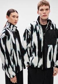 Fekete-fehér polárdzseki cikk-cakk mintával, álló gallérral és elöl cipzárral. Fekete díszítések az ujjakon és a zsebeknél.