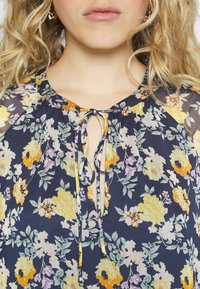 Blusa floral em azul marinho com padrões amarelos, lavanda e verdes. Apresenta um decote redondo com um fecho de laço e tecido texturizado.