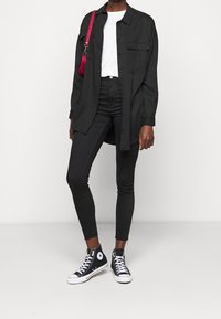 Zwarte button-up shirt over een witte t-shirt, gecombineerd met zwarte skinny jeans en zwarte high-top sneakers met witte accenten.