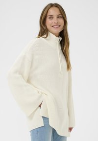 Jeune femme souriante, portant un pull en tricot crème avec une fermeture éclair semi-zip et des manches longues, associé à un jean bleu clair, se tenant devant un fond uni.