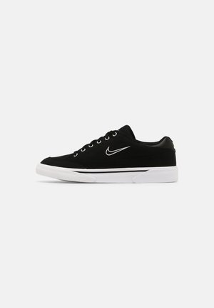 Czarny niski sneaker z białą podeszwą, białym logo Nike Swoosh i sznurowadłami na gładkim jasnoszarym tle.