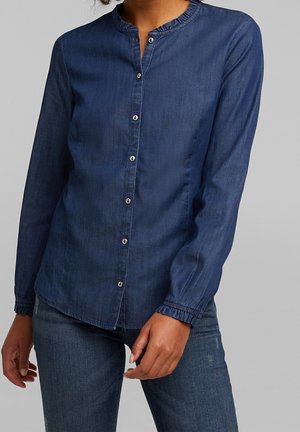Kvinde iført en langærmet denim skjorte med knapper og flæsekrave samt matchende denim jeans mod en ensfarvet baggrund.