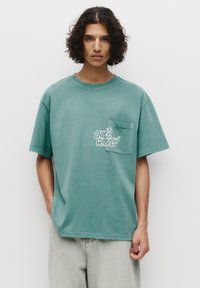 Grøn oversized t-shirt lavet af blød bomuld, med en brystlomme og et trykt logo i hvid tekst med flamme detaljer.