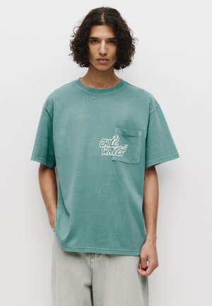 Grøn oversized t-shirt lavet af blød bomuld, med en brystlomme og et trykt logo i hvid tekst med flamme detaljer.