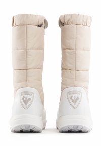 Beige geïsoleerde laarzen met een quilted ontwerp, een ronde bovenkant en een witte zool; logo gecodeerd op de hak; zichtbare ritssluiting details.