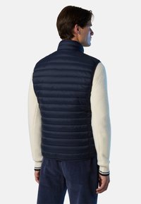 Mann, der sich abwendet, trägt eine marineblaue Steppweste über einem cremefarbenen Strickpullover und dunkelblauen Cordhosen vor einem einfarbigen Hintergrund.