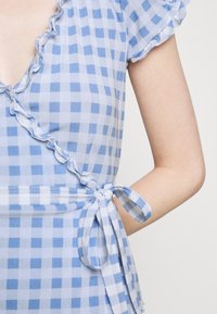 Abito avvolgente a scacchi blu e bianchi con scollatura a volant e maniche corte, legato in vita, che mostra parte del braccio di una persona.
