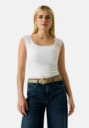 Donna bionda che indossa una canotta bianca in maglia, jeans blu scuro e una cintura beige, in piedi contro uno sfondo bianco semplice.