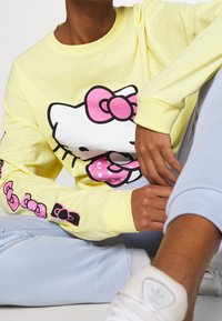 Camisa amarilla de manga larga con un gran diseño de Hello Kitty con un lazo rosa, adornada con colores pastel y pequeños gráficos de lazos en las mangas.