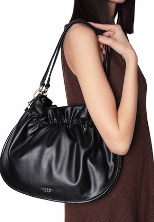 Donna con vestito marrone senza maniche che tiene una borsa a tracolla in pelle nera con dettagli arricciati e accessori dorati.