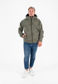 Olijfgroen jack met capuchon, meerdere voorkruiszakken, ritssluiting en een gestructureerde afwerking. Lichtblauwe jeans en witte sneakers.