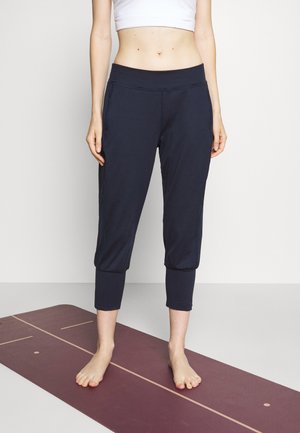 Barfodet person iført sorte, korte leggings og en hvid crop top, stående på en bordeaux yogamåtte i et studie.