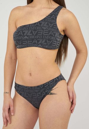 Vrouw draagt donkergrijze ééngeschouderde bikini met herhalend Emporio Armani EA7-logo patroon, toont het middenstuk en rechterhand rustend op de heup.