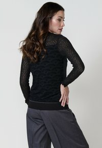 Un modello presenta un maglione nero, con un motivo intricatamente lavorato e maniche lunghe, abbinato a pantaloni grigi a gamba larga, in piedi contro uno sfondo neutro.