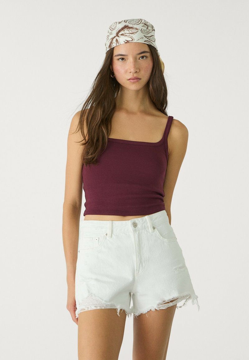Burgunderfarbener, taillierter Tanktop mit Spaghetti-Trägern, kombiniert mit weißen, abgeranzten Denim-Shorts mit fransigen Kanten. Kopftuch mit gemusterten Akzenten.