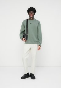 Green sweatshirt with a small black logo, paired with cream trousers and black sneakers. Carrying a small black bag across the body.