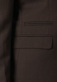 Veste de costume marron foncé texturée avec deux poches horizontales, une poche à rabat en bas et une poche passepoilée en haut.