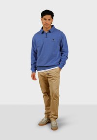 Pullover blu con mezza zip, dotato di un piccolo logo. Abbinato a pantaloni khaki e sneaker grigie con lacci bianchi e suole marroni.