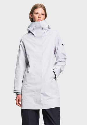 Personne portant un imperméable à capuche gris clair avec un col montant et les mains partiellement dans les poches, debout devant un fond uni.