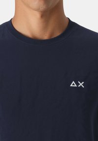 Uomo che indossa una maglietta girocollo blu navy con triangoli e simboli X ricamati bianchi sul petto.
