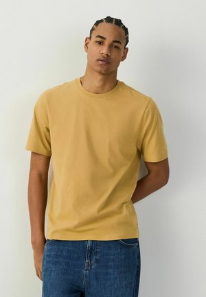 Ung mand med fletninger iført en sennepsgul kortærmet t-shirt og blå jeans, stående mod en ensfarvet lys væg.