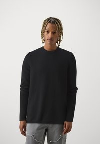 Suéter negro texturizado con cuello redondo y mangas largas. Combinado con jeans grises desgastados, que presentan costuras visibles y bolsillos.