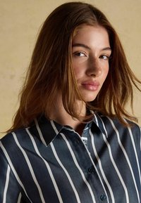 Tume sinine ja valge triibuline nööbiga särg, millel on krae, sile kangastekstuuri ja kontrastsete nupudega.