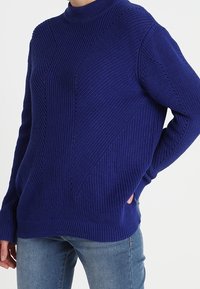 Modrý pletený svetr s vysokým krkem, zdobený texturovanými vzory a bočními kapsami, v kombinaci s džínovými kalhotami. Uvolněný střih.