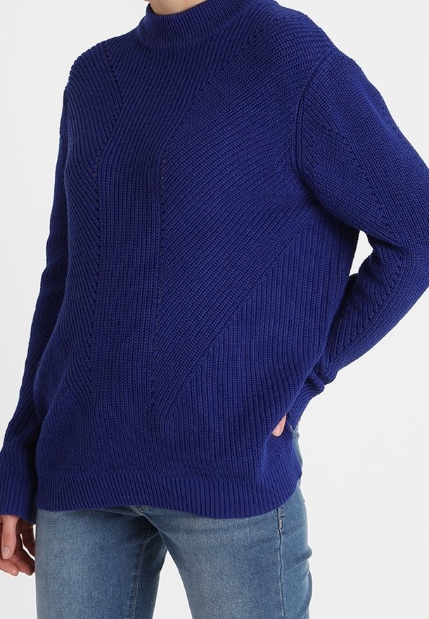 Modrý pletený svetr s vysokým krkem, zdobený texturovanými vzory a bočními kapsami, v kombinaci s džínovými kalhotami. Uvolněný střih.