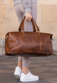 Smeđi kožni sportski kofer s dva gornja ručka, prednjim zatvaračem i srebrnim metalnim detaljima. Glatka tekstura s vidljivim šavovima.