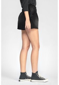 Osoba oblečená v černých denímových šortkách a černých vysokých teniskách s bílými tkaničkami, stojící proti jednoduchému světlému pozadí.