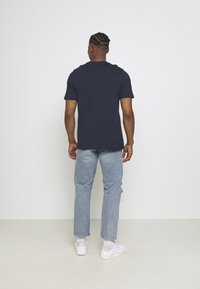 Marinblå bomullströja med korta ärmar, kombinerad med ljusblå slitna jeans och vita sneakers, sedd bakifrån.