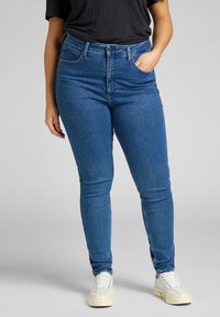 Jeans typu wysokiej talii, prostą nogawką w niebieskim denimu o gładkiej fakturze, z pięcioma kieszeniami i zapięciem na guzik. Białe sneakersy dopełniają stylizację.