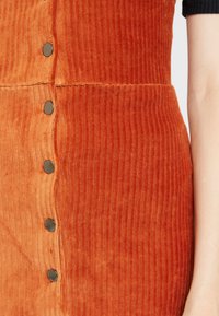 Abito in velluto a coste arancione bruciato con righe verticali, dotato di cinque bottoni a pressione metallici lungo la cucitura laterale per la chiusura.