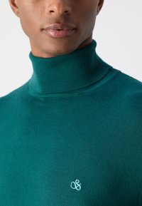Pull à col roulé teal avec un col texturé et côtelé, et un petit logo brodé en bleu clair sur la poitrine.