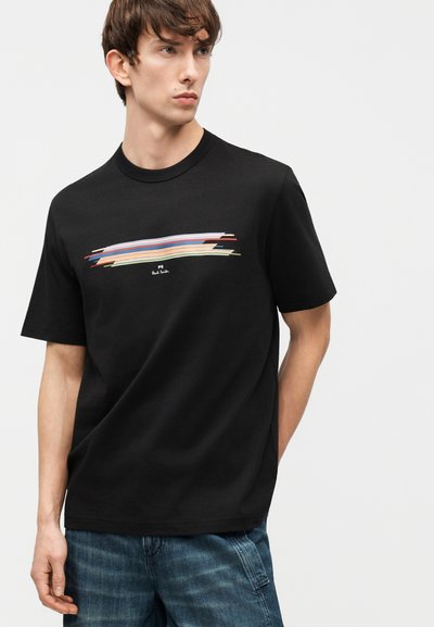 Νεαρός άνδρας που φοράει μαύρο T-shirt με πολύχρωμο οριζόντιο μοτίβο από πινελιές και σκούρο μπλε τζιν, σε απλό φόντο.