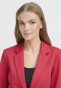 Blazer rouge avec un design structuré, col à revers et une coupe ajustée. Le modèle porte un haut noir en dessous. Texture du tissu lisse.