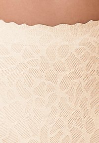 Close-up van beige kantstof met bloemmotieven tegen een lichte huid achtergrond.