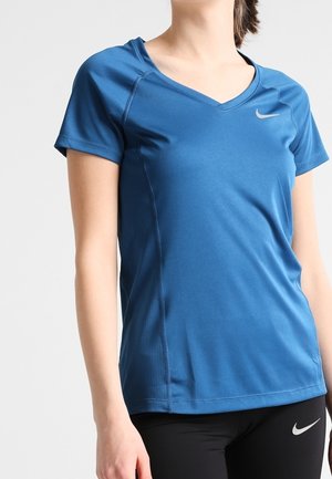 Kobieta ubrana w niebieską sportową koszulkę Nike z krótkim rękawem i dekoltem w serek oraz czarne legginsy z widocznym logo Nike na obu elementach garderoby.