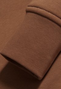 Tela marrón con una textura fina, que muestra el detalle de un puño doblado. El material tiene una apariencia suave y ligera, con un grosor sutil.
