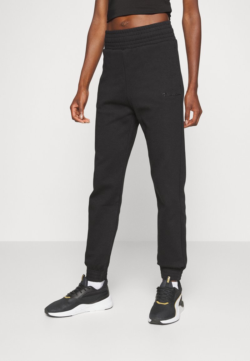 Pantalones de chándal negros con cinturilla elástica acanalada y puños en los tobillos. Presenta un detalle de logo en el lado izquierdo y una textura suave.