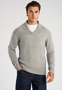 Grå stickad tröja med sjalkrage, långa ärmar och ribbstickade muddar. Buren över en vit t-shirt, kombinerad med mörkblå jeans.