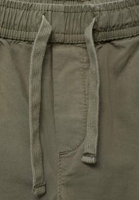 Närbild av en olivgrön elastisk midjeband med två hängande tygsnören på avslappnade byxor eller shorts.