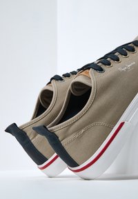 Baskets en toile beige avec lacets bleu marine, semelle en caoutchouc et accent rouge. Présente un patch logo en cuir beige sur le côté et une semelle texturée.