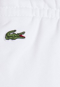 Bílá látka s malým zeleným a červeným vyšívaným logem krokodýla. Textura vypadá hladce s jemným leskem.