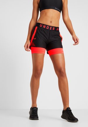 Sportlerin trägt schwarzen Sport-BH, schwarze und rote Under Armour Shorts mit roten inneren Leggings und schwarze Adidas Laufschuhe, steht auf grauem Boden.