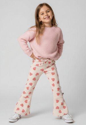 Uśmiechnięta dziewczyna stoi z rękami na biodrach, ubrana w różowy sweter, białe rozszerzane spodnie w czerwone serca oraz białe tenisówki na jednolitym szarym tle.