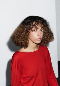 Pull rouge à manches longues avec un large décolleté, une texture douce et une coupe décontractée ; le modèle a des cheveux bouclés jusqu'aux épaules et un arrière-plan neutre.